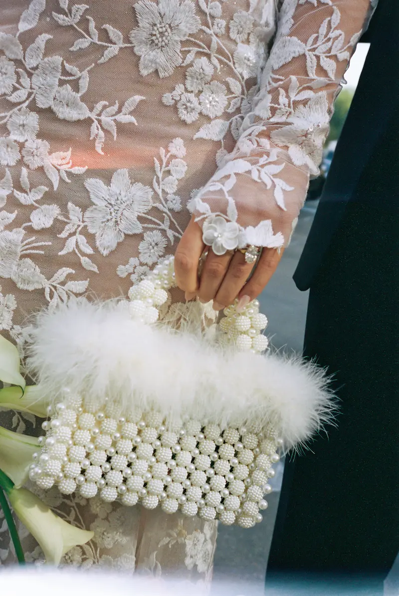 Detail: Perlentasche mit Federn & Ring Nahaufnahme einer eleganten weißen Perlentasche mit Federn, getragen zur floralen Spitze – Hochzeitsaccessoires mit Charakter.